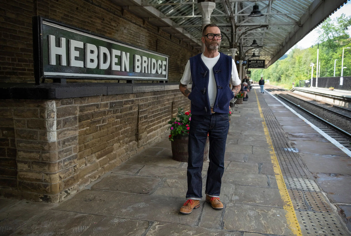 A man wearing glasses, a navy vest over a white t-shirt, and dark jeans stands on a train station platform next to a brick wall with a 'HEBDEN BRIDGE' sign.