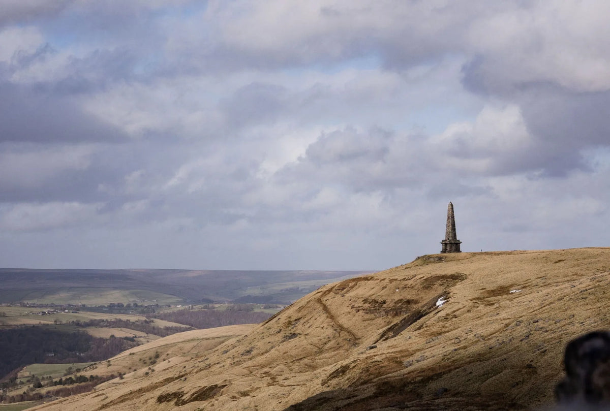A wide landscape photograph of a grassy brown hillside under a cloudy blue sky, with a small monument or obelisk visible near the top right and a distant valley below.