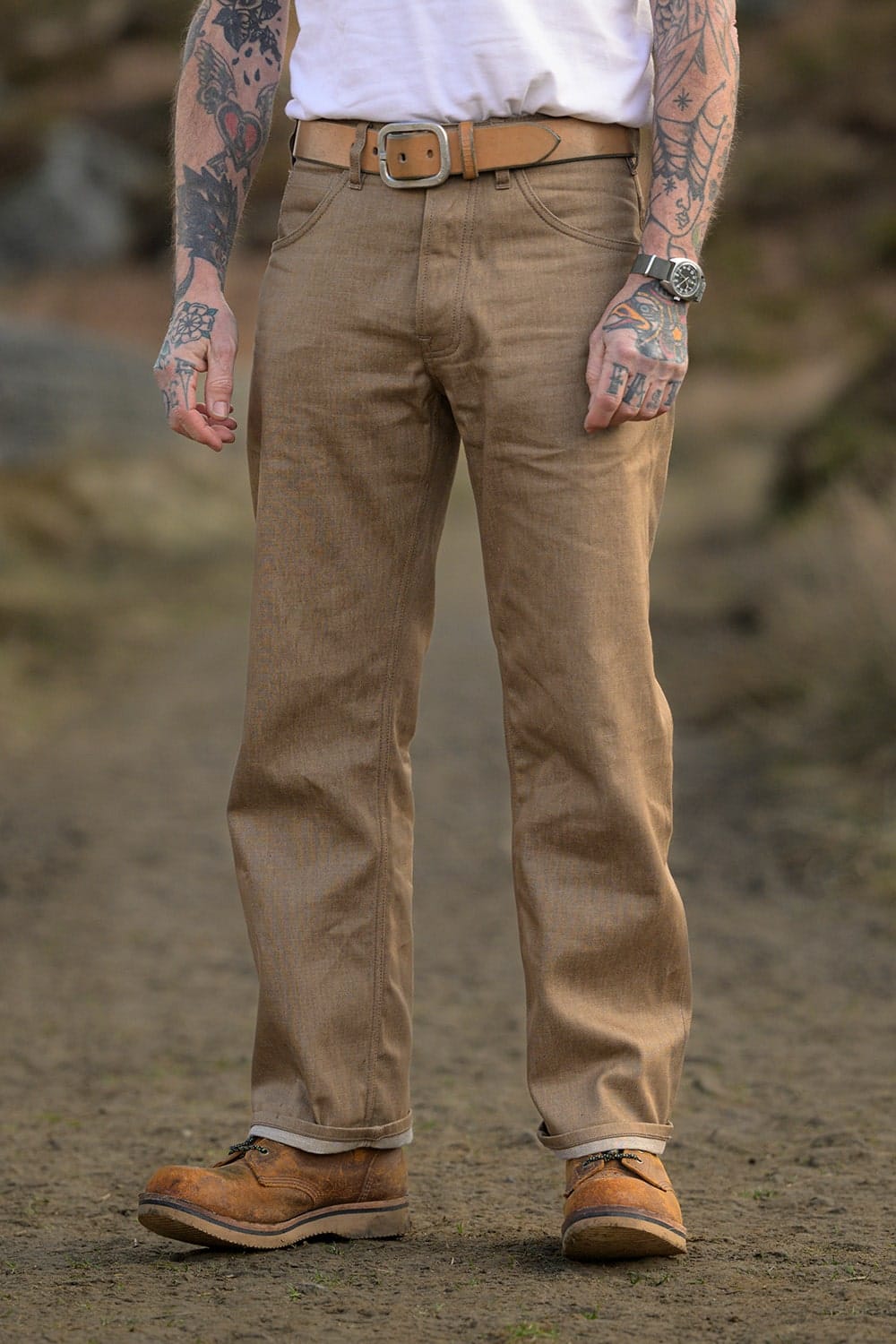 A person standing outdoors wearing tan straight-leg work pants with a brown belt, a white shirt, and brown work boots. The background is a blurred natural outdoor setting.