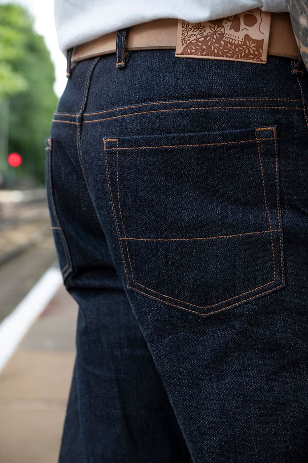 Close-up back view of a person wearing dark indigo denim jeans with contrast tan stitching, a tan leather belt, and a skull patch on the back waistband.
