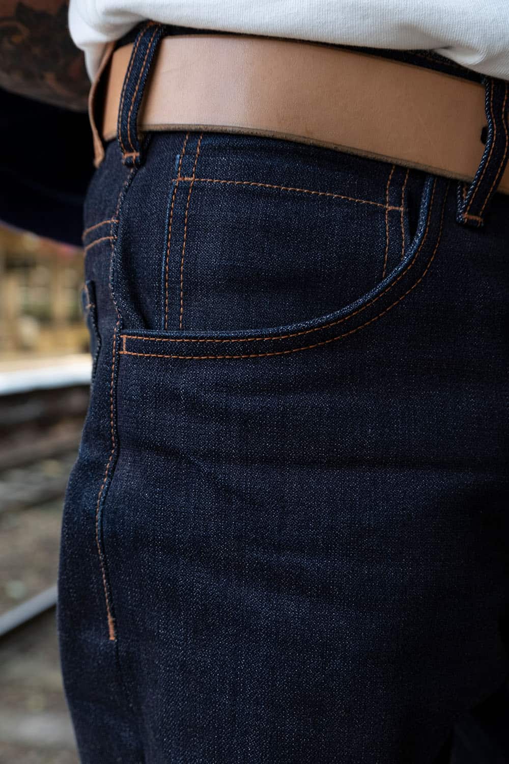 Close-up of dark indigo denim jeans with contrast orange stitching, worn with a tan leather belt and white shirt.