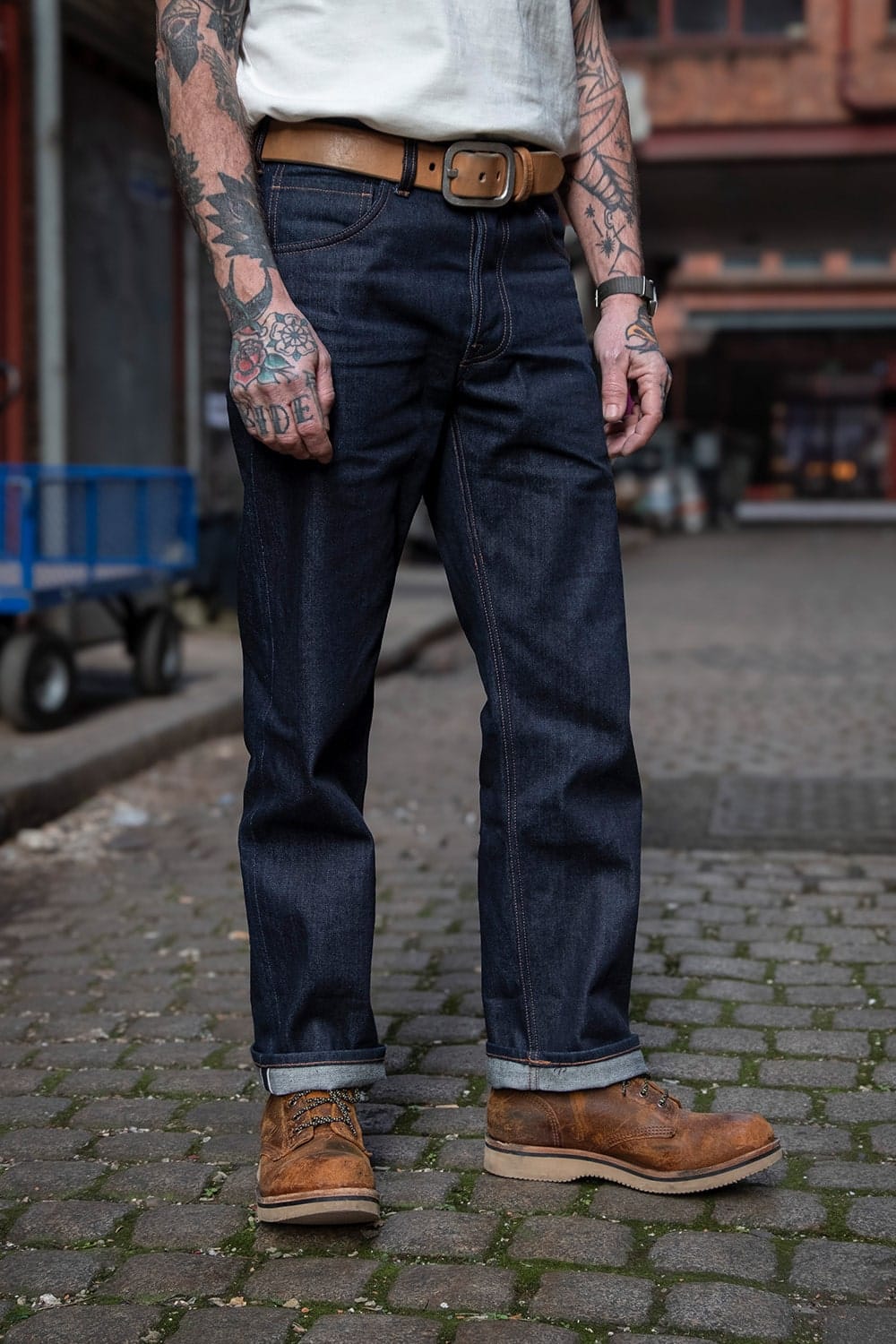 A person standing on a cobblestone street wearing dark blue denim jeans with a tan leather belt, a white shirt, and brown work boots.