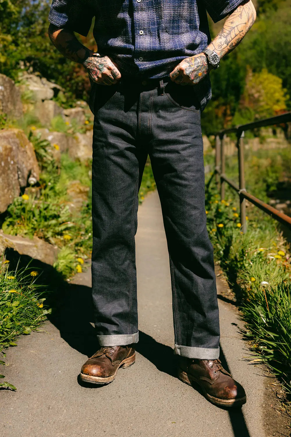 A person standing on an outdoor paved path wearing dark charcoal gray cuffed jeans, a blue plaid button-up shirt, and brown leather shoes.