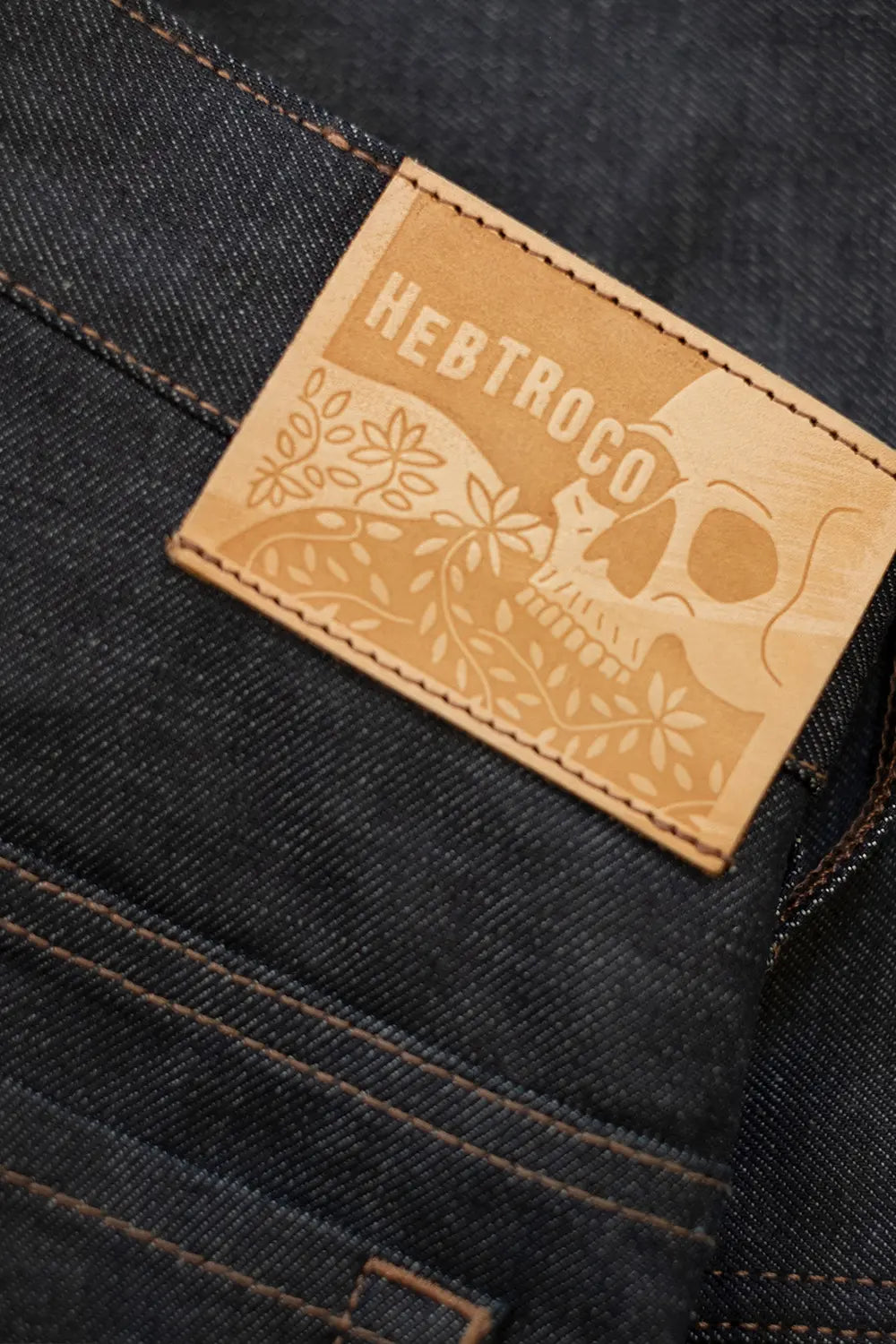 Close-up of dark indigo denim jeans showing a tan leather patch with the brand name 'HEBTROCO' and a floral skull design.