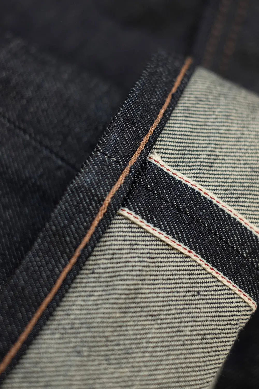Close-up detail of dark indigo denim fabric showing the contrast between the dark indigo outer side and the lighter gray ribbed inner side with tan stitching.