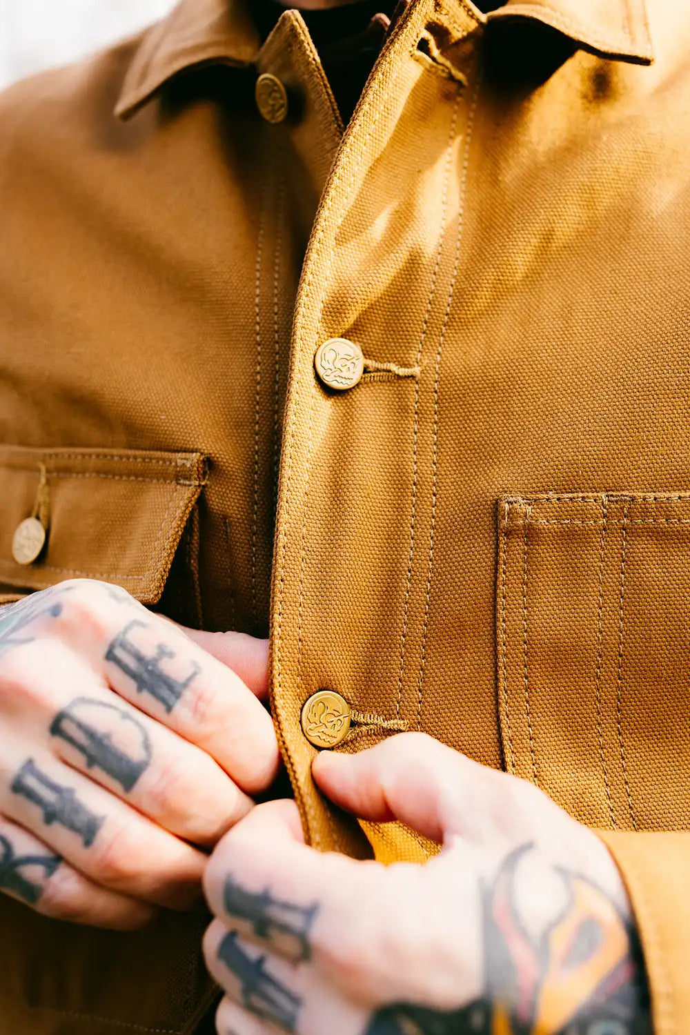 Close-up of a person wearing a bob job tan chore coat and fastening the front button with both hands, which have black tattoos.