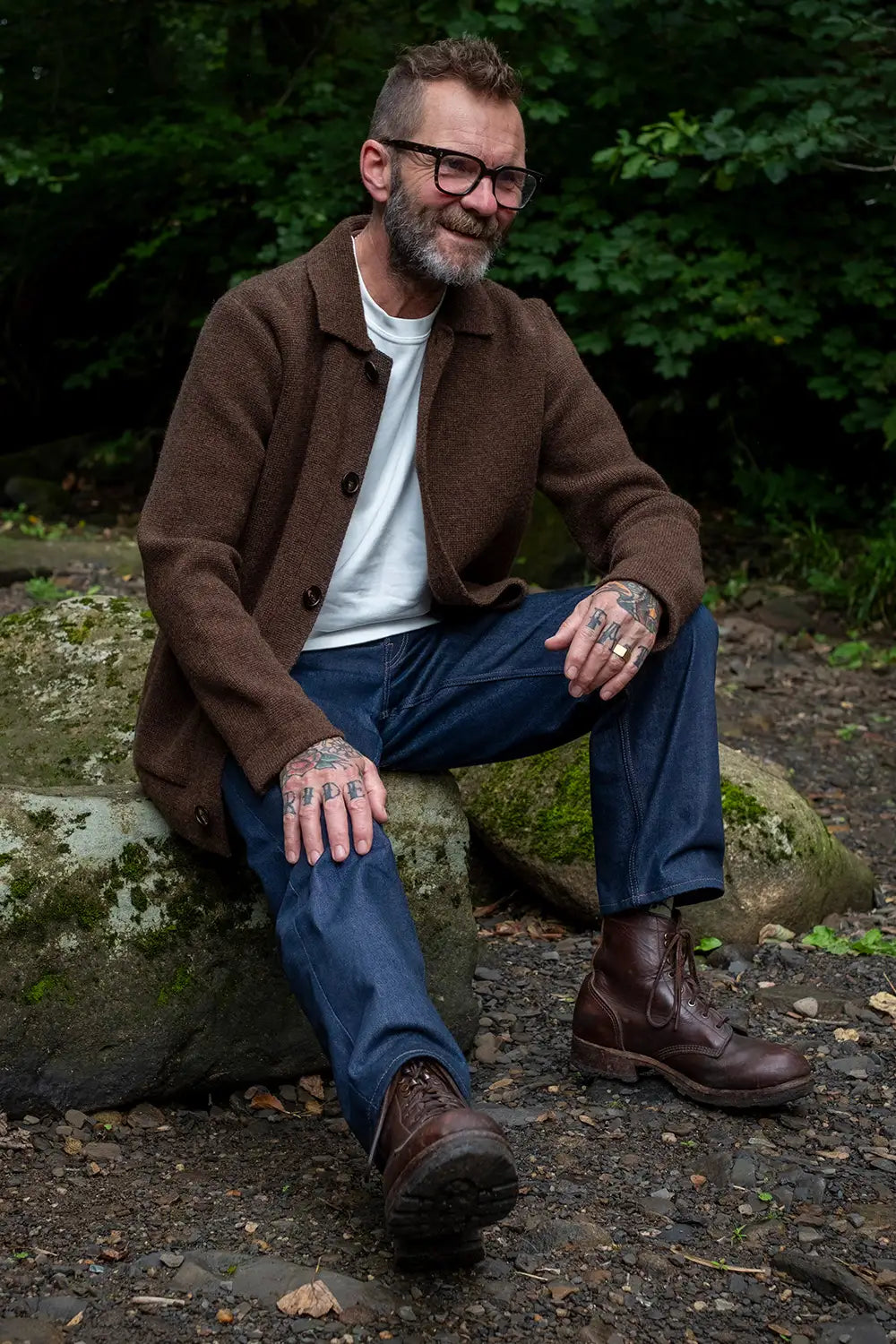 A man with glasses, a beard, and tattoos sits on a mossy rock outdoors in a wooded setting. He is wearing a brown button-up cardigan over a white t-shirt, blue jeans, and brown leather boots.