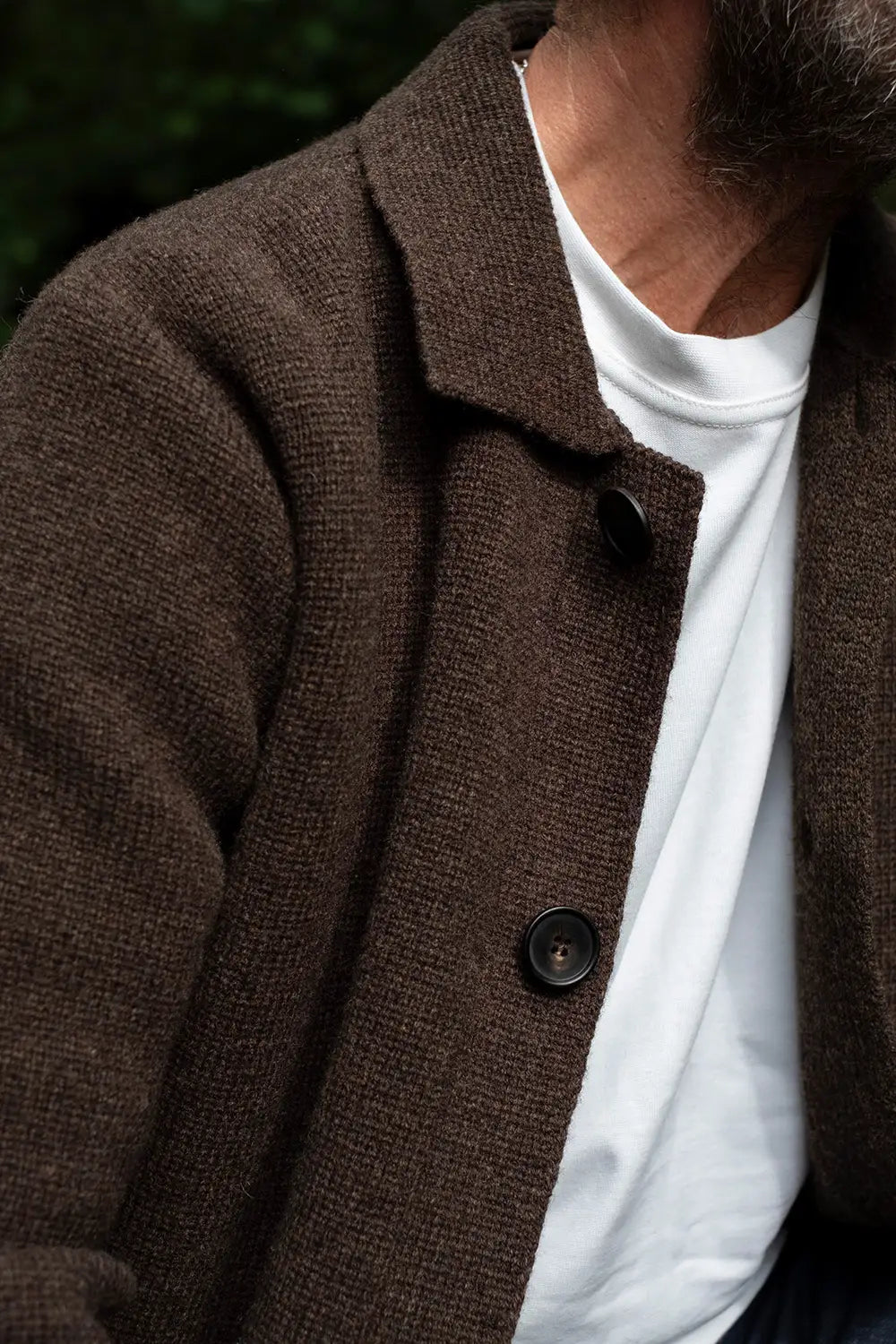 A close-up of a person wearing a brown textured knit cardigan over a white t-shirt, with a dark button and a blurred outdoor background.