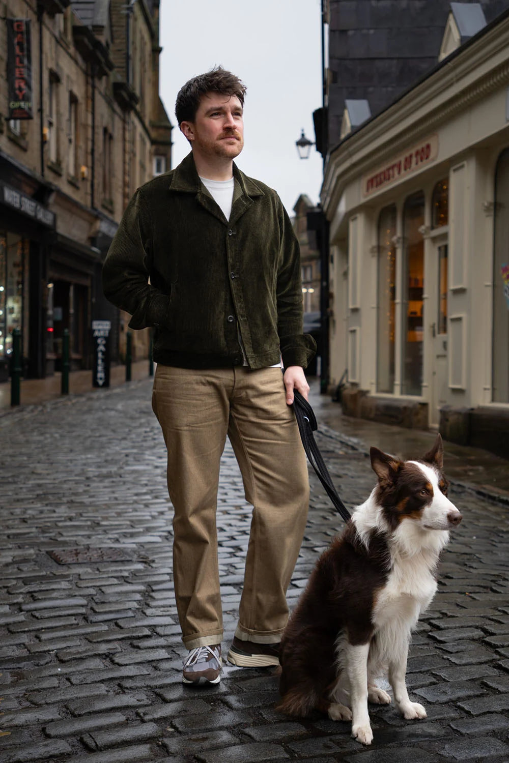 A man wearing a dark olive green corduroy jacket, beige trousers, and sneakers walks a brown and white Border Collie on a cobblestone street in an urban setting.