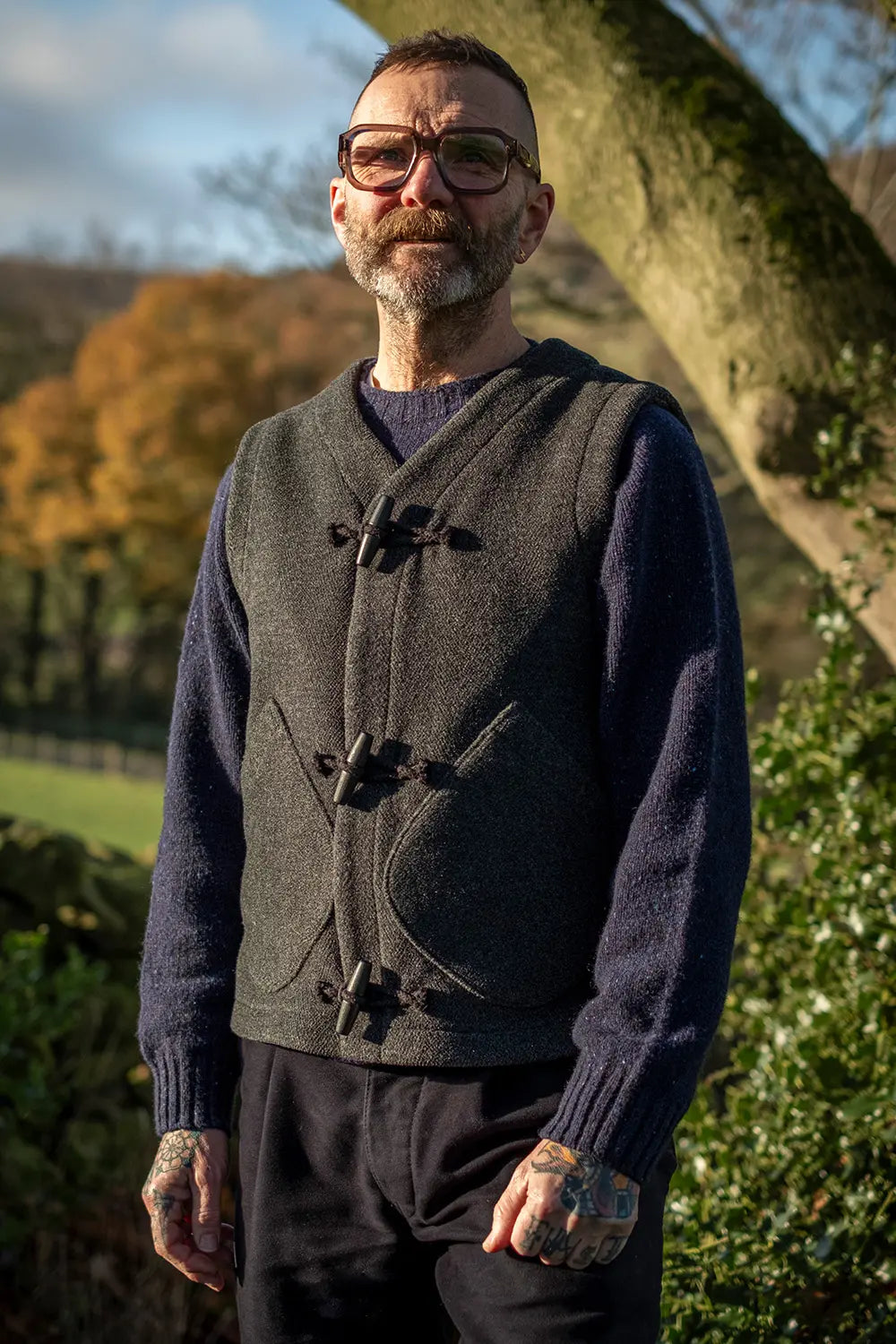 A man wearing a dark gray wool teasel duffel vest over a navy blue knit jumper, standing outdoors in front of a tree with autumn foliage in the background.