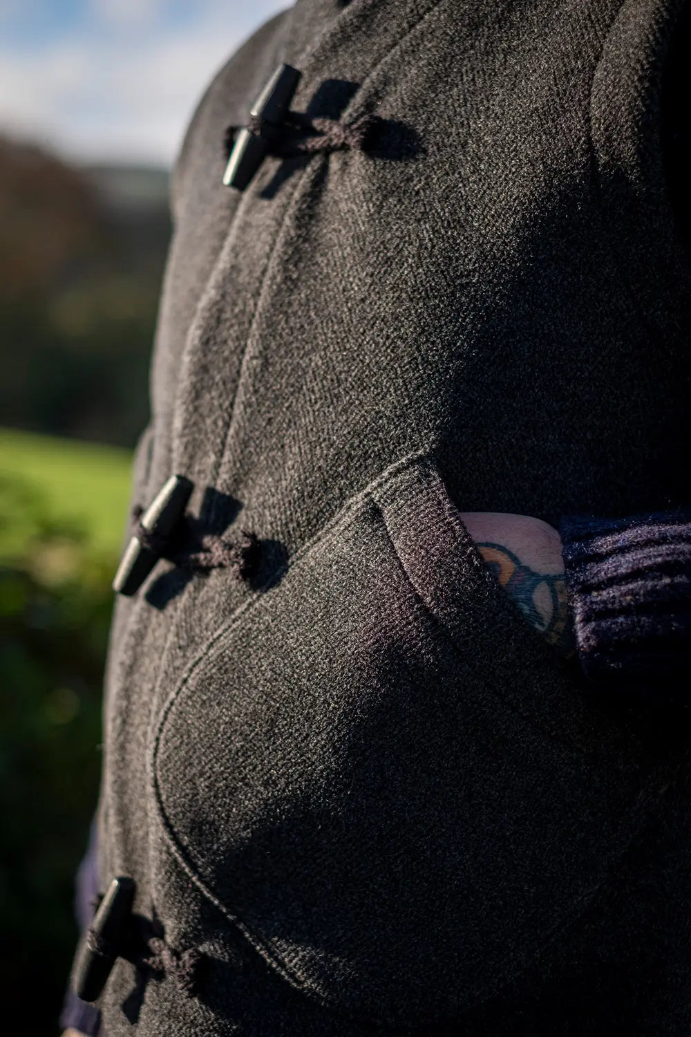 Close-up of a dark charcoal gray wool duffel coat with black toggle closures, worn outdoors with a purple knit sleeve visible in the pocket.