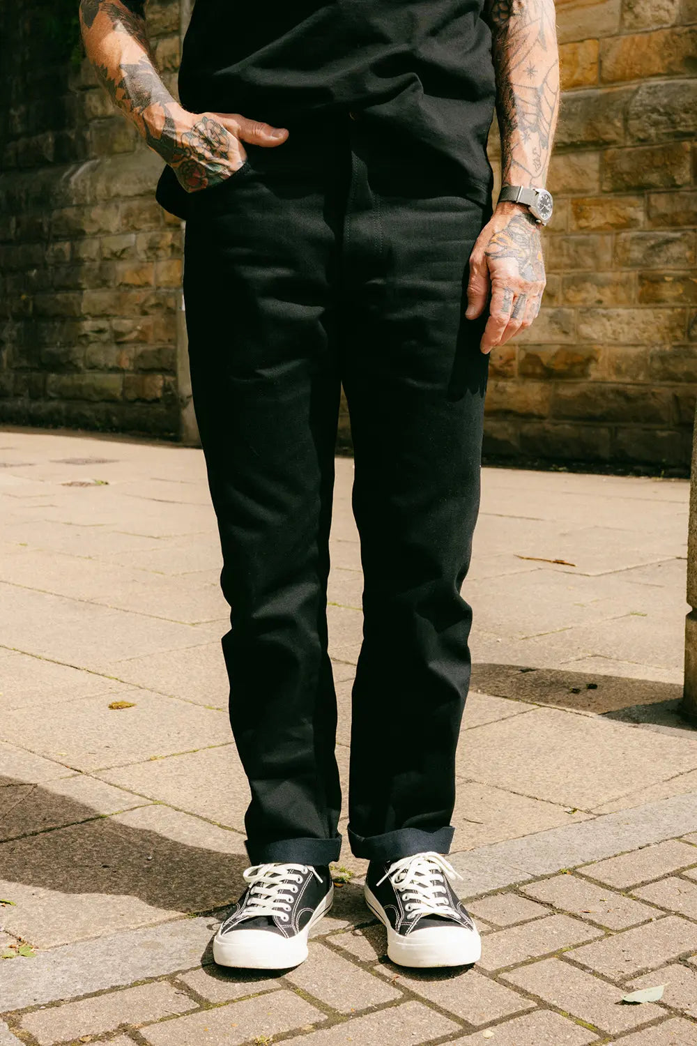 A person wearing black slim-fit trousers, a black t-shirt, and black and white sneakers, standing outdoors on a paved stone surface with a brick wall in the background.