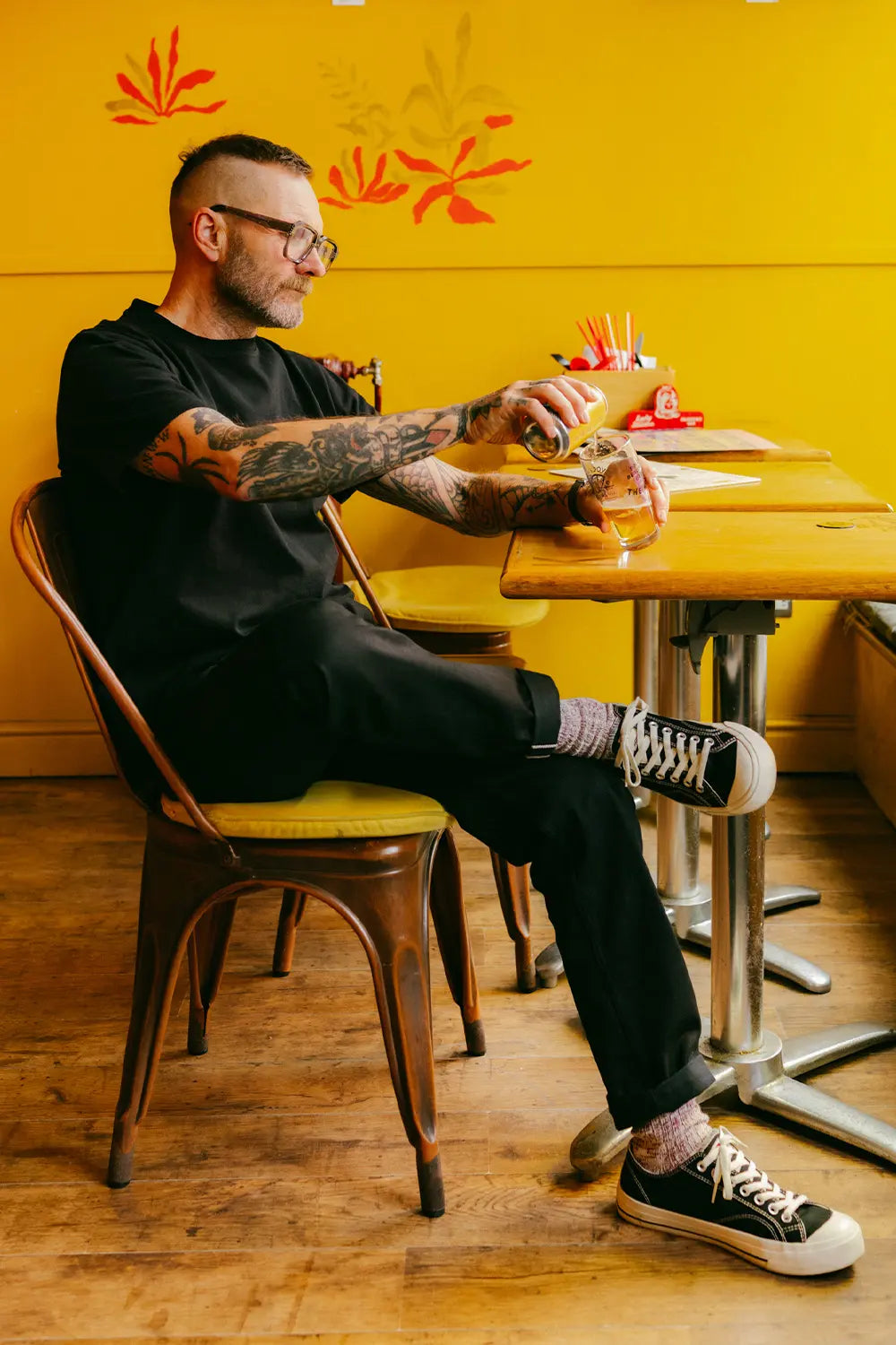 A man with tattoos and glasses sits in a wooden chair at a cafe table, wearing a black t-shirt, black pants, and black canvas sneakers against a bright yellow wall.