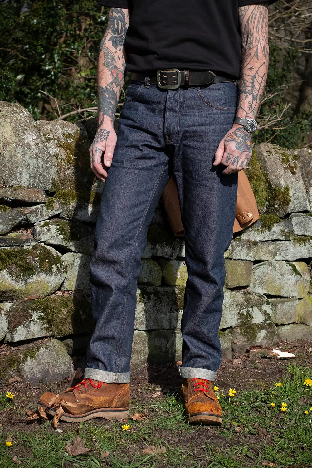 A person outdoors wearing dark blue denim jeans, a black t-shirt, a black belt, and brown leather boots, standing in front of a mossy stone wall.