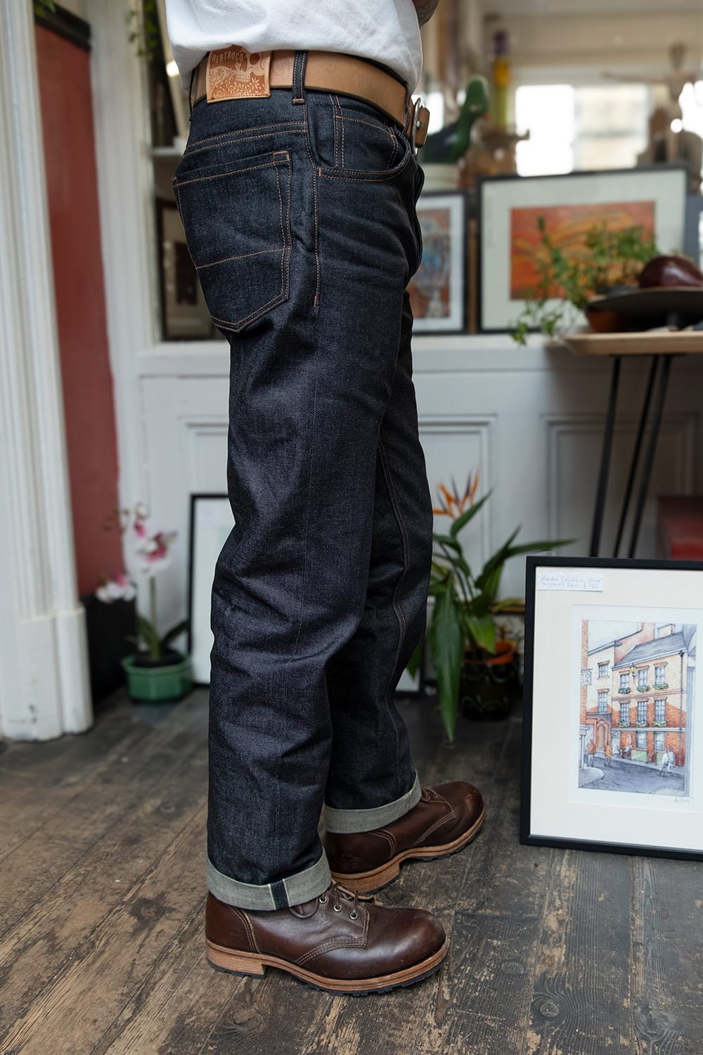 A person shown from the waist down wearing dark indigo denim jeans with a brown leather belt and brown leather lace-up boots, standing in a styled indoor room with framed art and plants.