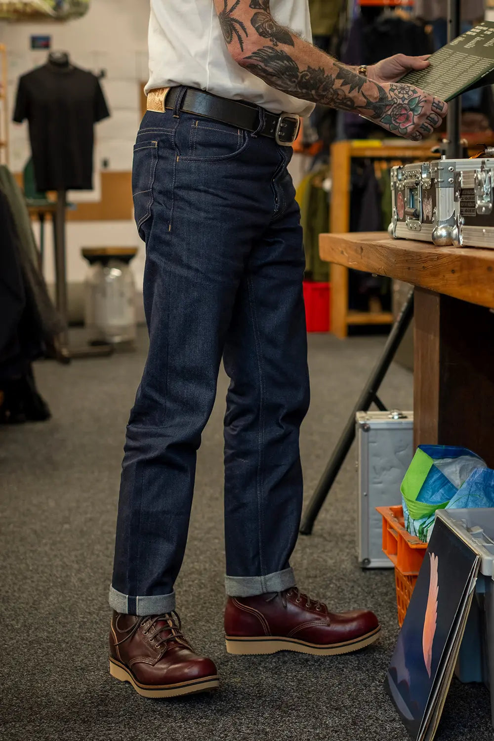 A person wearing dark blue denim jeans with rolled cuffs, a white shirt, and brown leather shoes, standing in a retail or workshop environment.