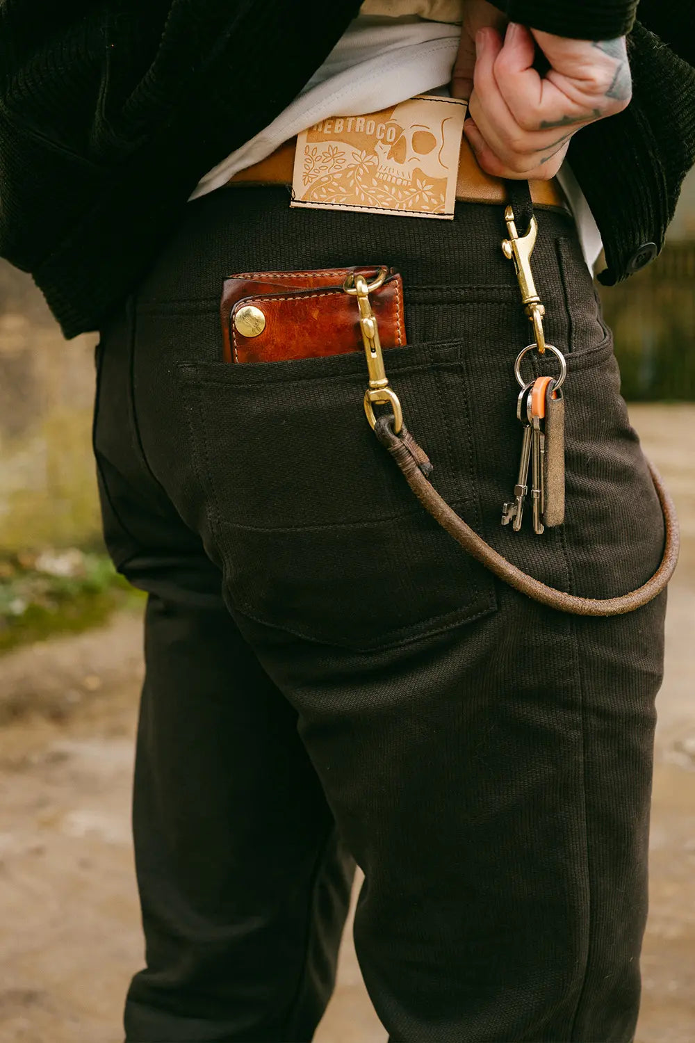 Close-up of a person wearing dark brown canvas pants with a brown leather belt, a brown leather utility pocket on the back pocket, and a leather keychain strap with brass hardware.