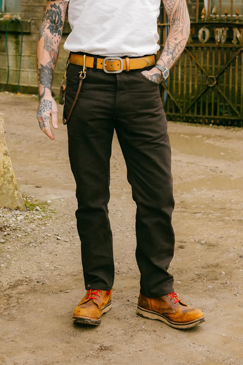 A person wearing dark brown work pants with a tan leather belt, a white sleeveless top, and brown leather work boots, standing outdoors on a dirt ground.