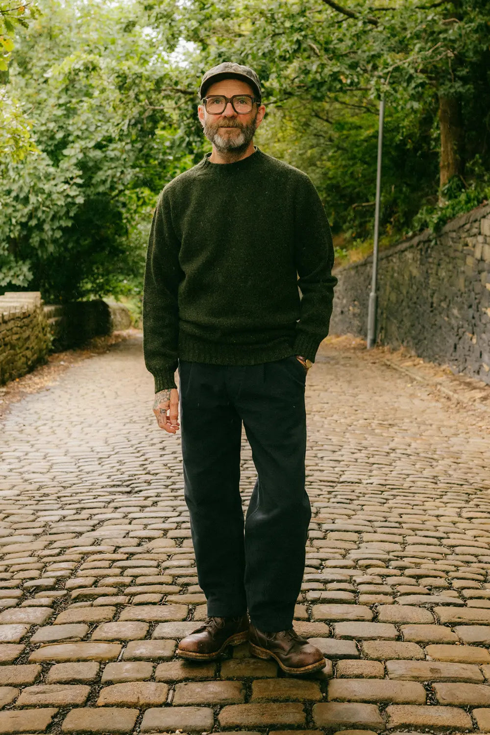 A man wearing a dark green knit sweater, dark trousers, brown leather shoes, and a cap stands on a cobblestone path surrounded by green trees and foliage.