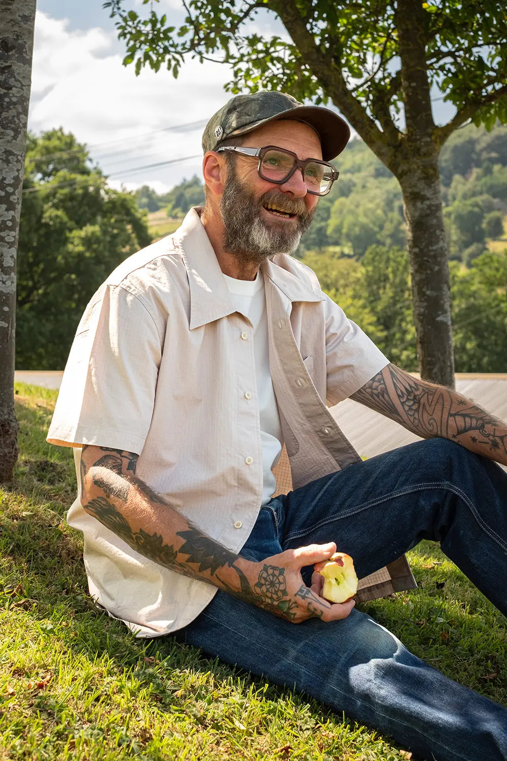 A smiling man with a beard, glasses, and tattoos sits on grass outdoors under trees, wearing a light beige short-sleeve button-up shirt over a white undershirt and blue jeans, while holding a sliced apple.