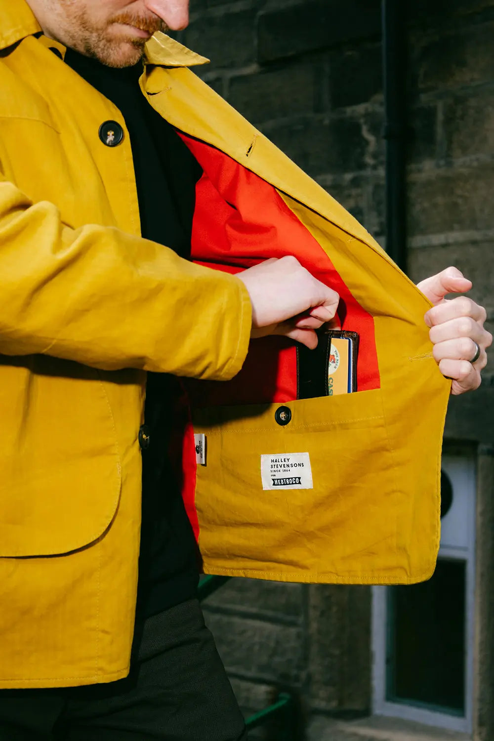 A close-up of a man wearing a mustard yellow waxed canvas jacket, opening the front to reveal a bright red interior lining and a front utility pocket holding cards.