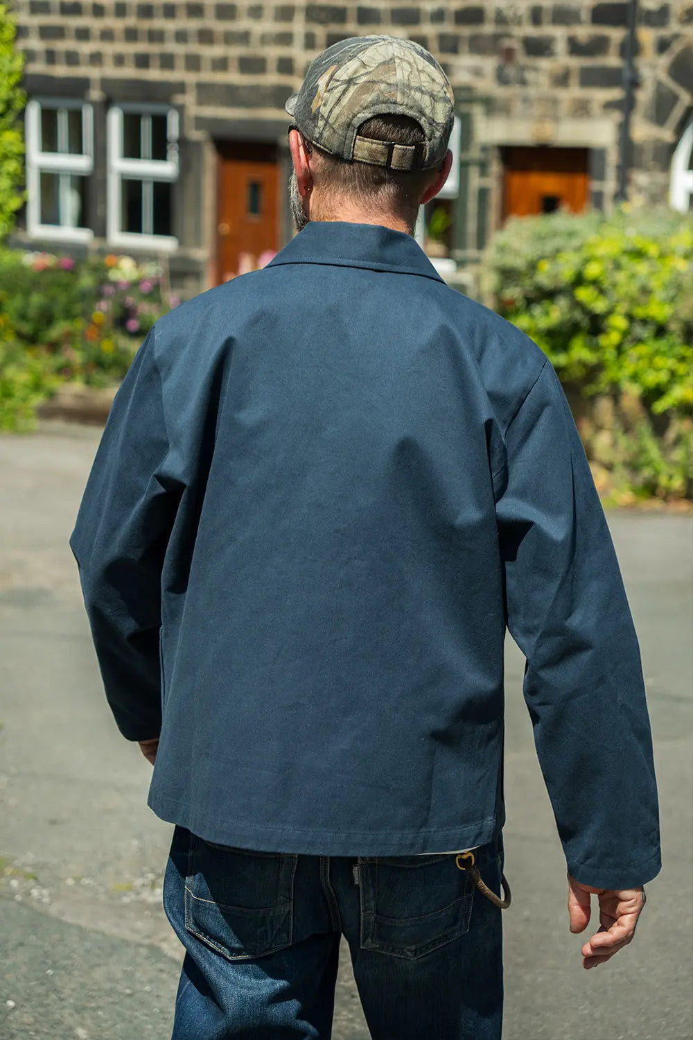 A man viewed from behind wearing a dark navy blue work jacket, blue jeans, and a camouflage baseball cap, standing outdoors in front of a stone building.