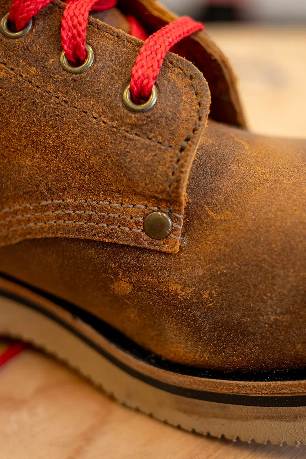 Close-up of a brown suede lace-up boot with bright red laces and brass eyelets, resting on a light wood surface.