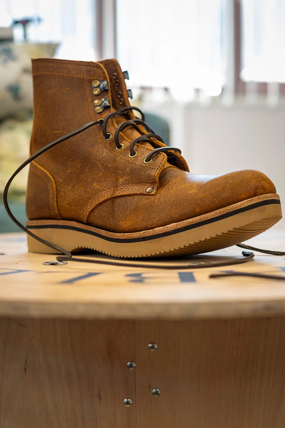 A single brown suede lace-up boot resting on a wooden surface in a softly blurred indoor setting.