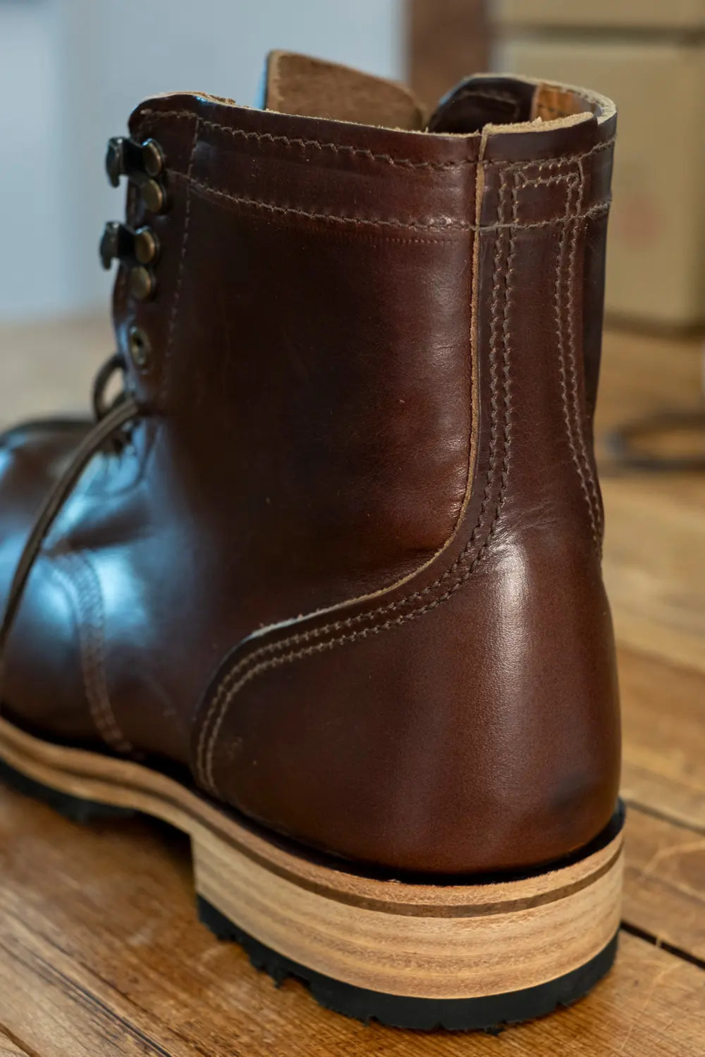 A close-up shot of a dark brown leather lace-up boot, showing the back and side details, resting on a wooden floor.