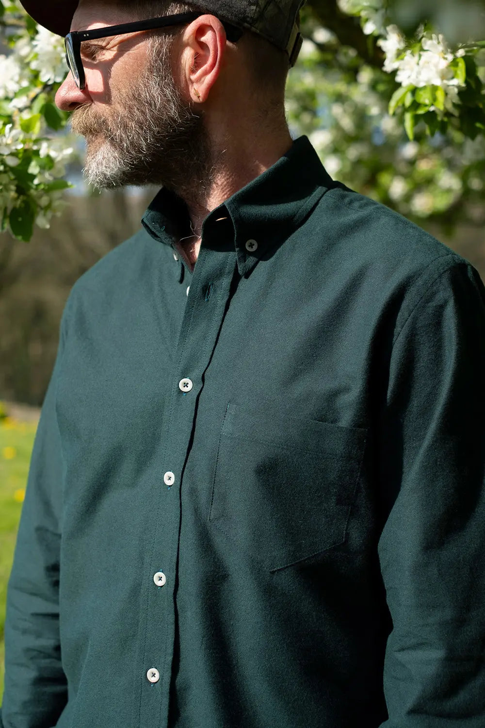 A man wearing a dark forest green long-sleeve button-down shirt, photographed outdoors in natural sunlight with blurred green foliage and white blossoms in the background.