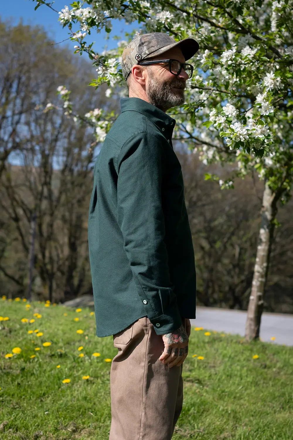 A man wearing a dark green long-sleeve button-up shirt, tan pants, and a camouflage cap stands in profile outdoors under flowering trees on a grassy hill.