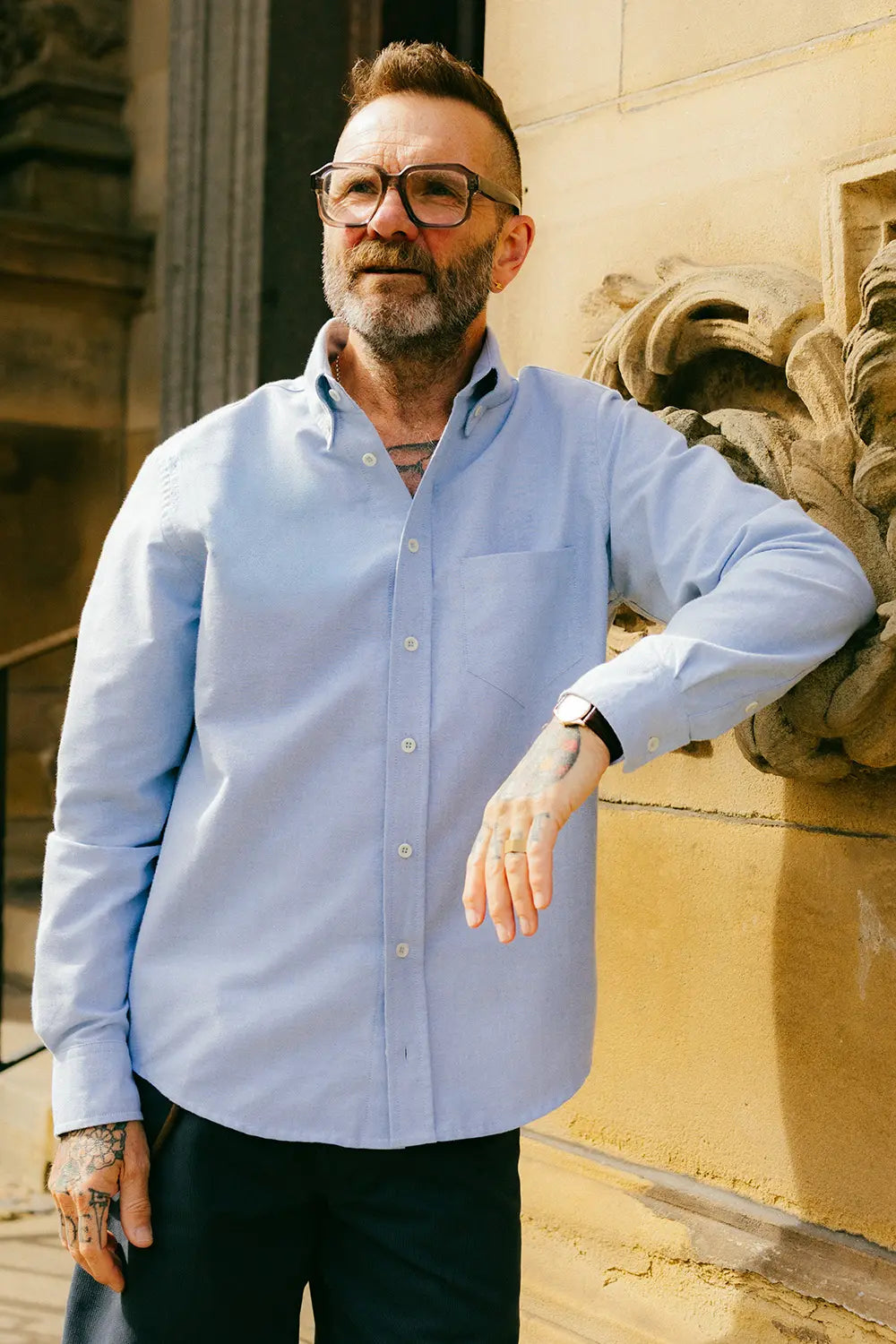 A man with glasses, a beard, and tattoos wearing a light blue long-sleeve button-down shirt and dark trousers, standing outdoors against a beige stone wall with classical carvings.