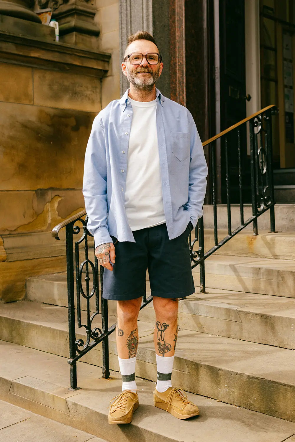 A man wearing glasses, a light blue button-down shirt over a white t-shirt, dark shorts, white socks, and tan sneakers, standing on stone steps in front of a building.