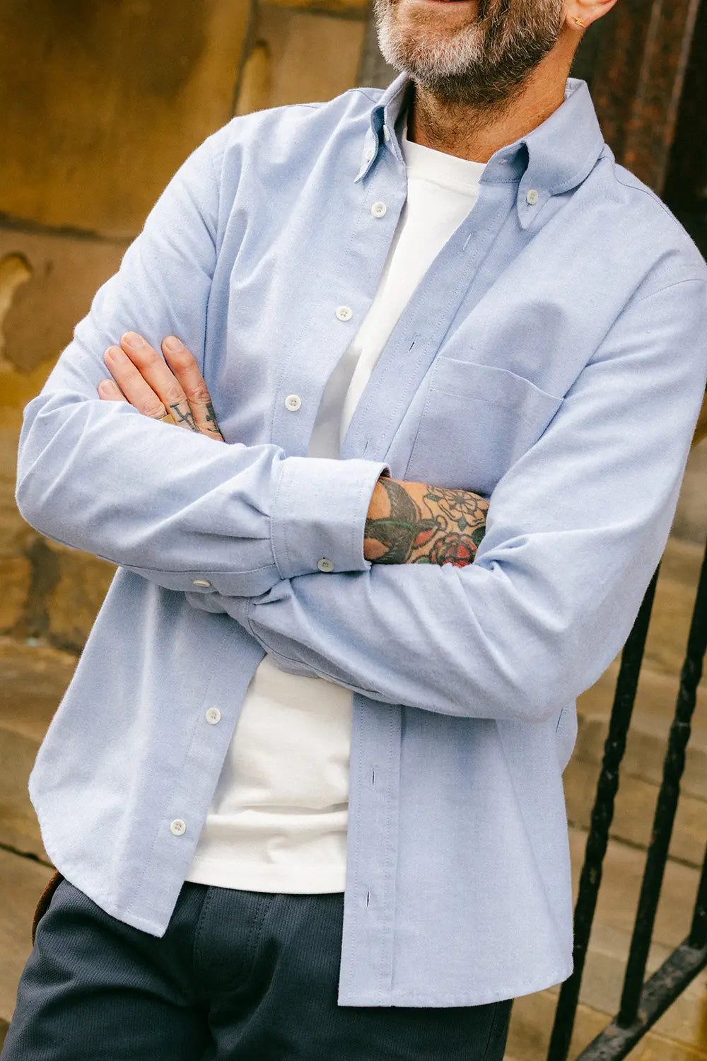 A man wearing a light blue long-sleeve button-down shirt over a white t-shirt and dark pants, standing with arms crossed.