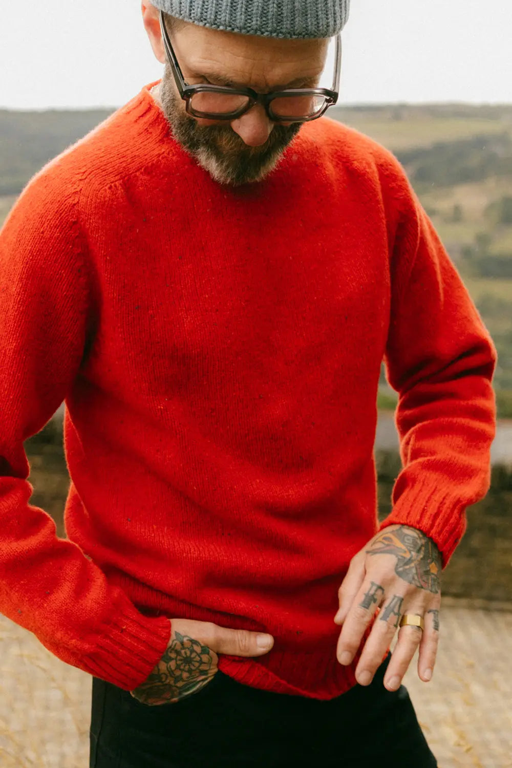 A man wearing a vibrant red knit jumper, black pants, a gray beanie, and glasses, standing outdoors with a blurred landscape background.