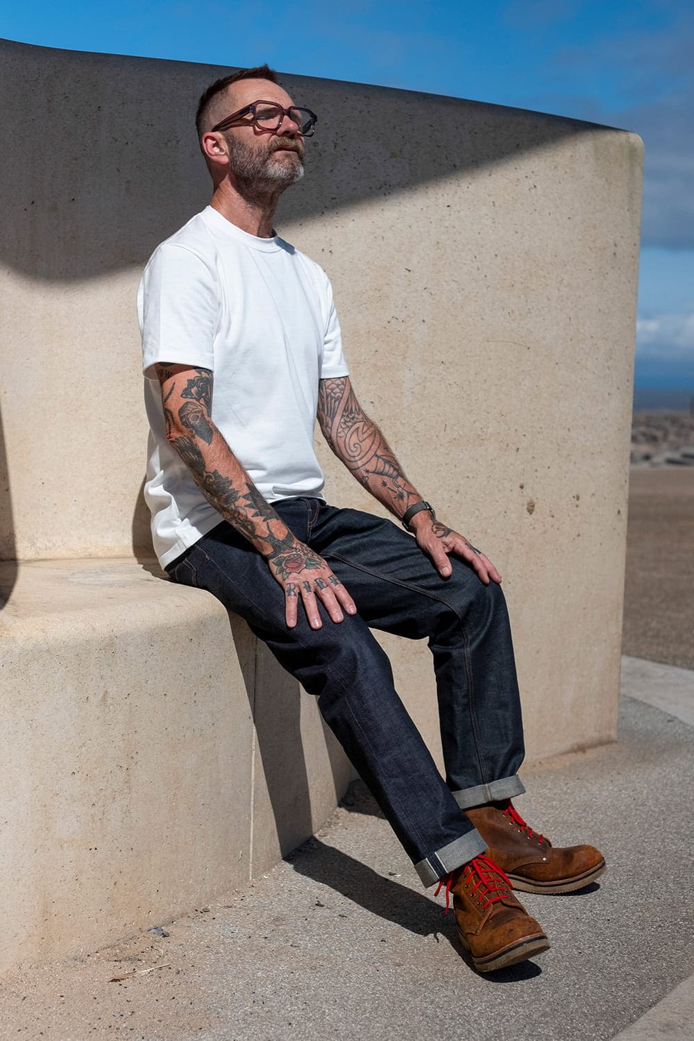 A man with tattoos and glasses sits on a concrete outdoor structure, wearing a white t-shirt, dark blue jeans, and brown boots with red laces.