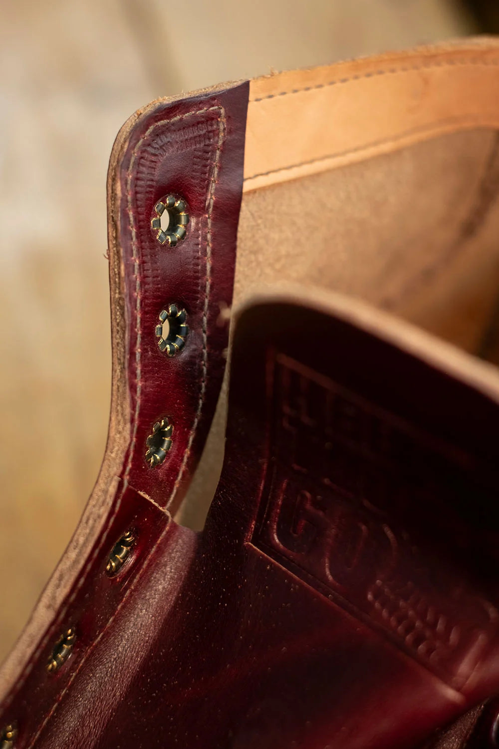 Close-up of a leather boot corner showing a deep burgundy outer leather panel with metal rivets, contrasted against a tan inner leather lining and embossed branding.