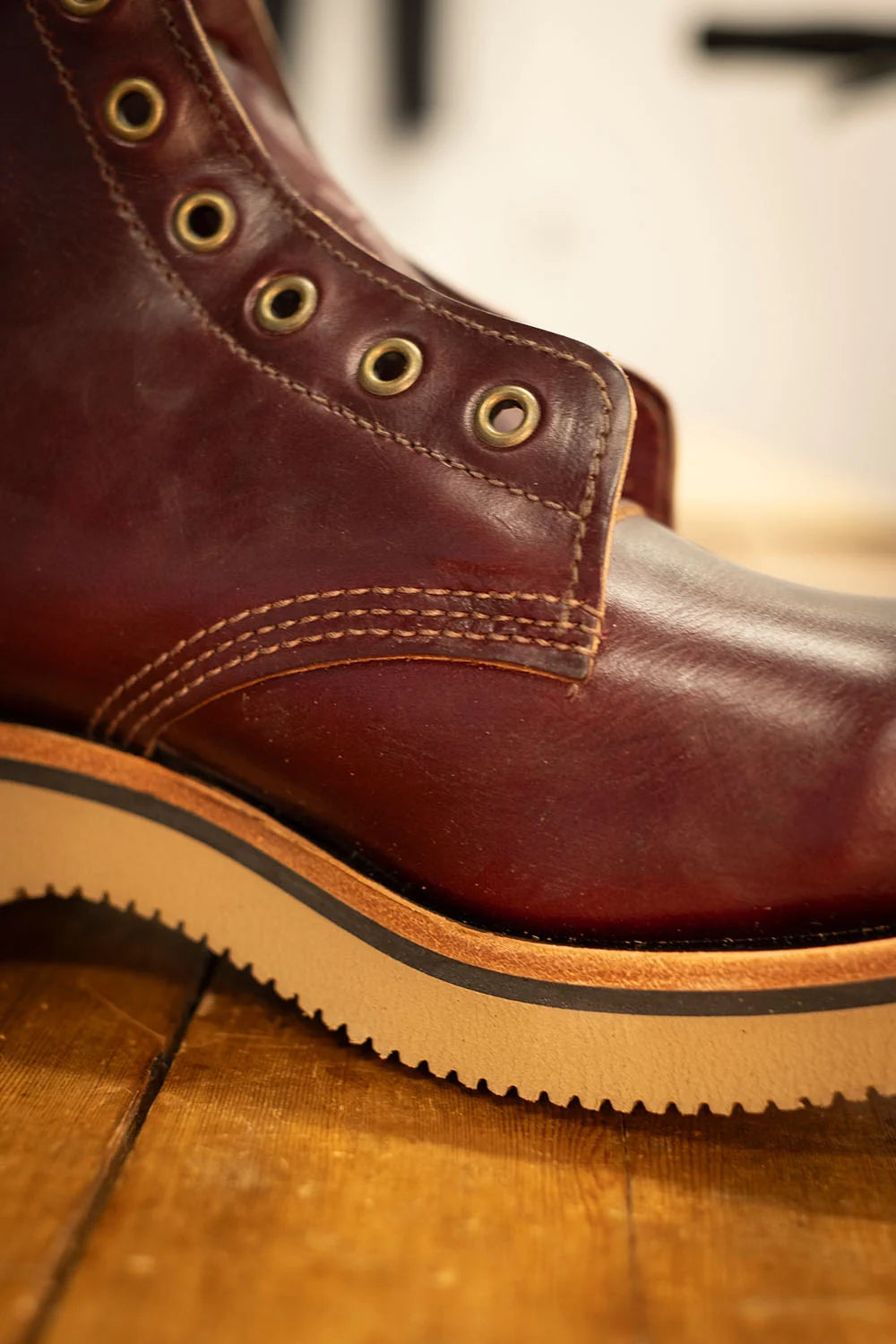 A close-up shot of a dark reddish-brown leather lace-up boot resting on a wooden floor, showing the textured sole and brass eyelets.