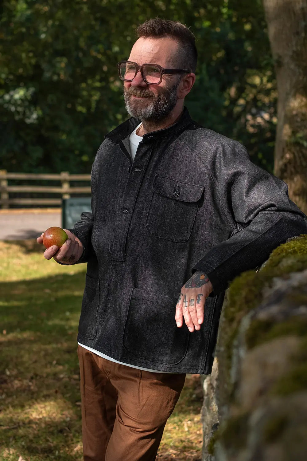 A man with a beard and glasses wearing a dark charcoal gray utility overshirt and brown trousers, leaning against a mossy stone wall outdoors while holding an apple.