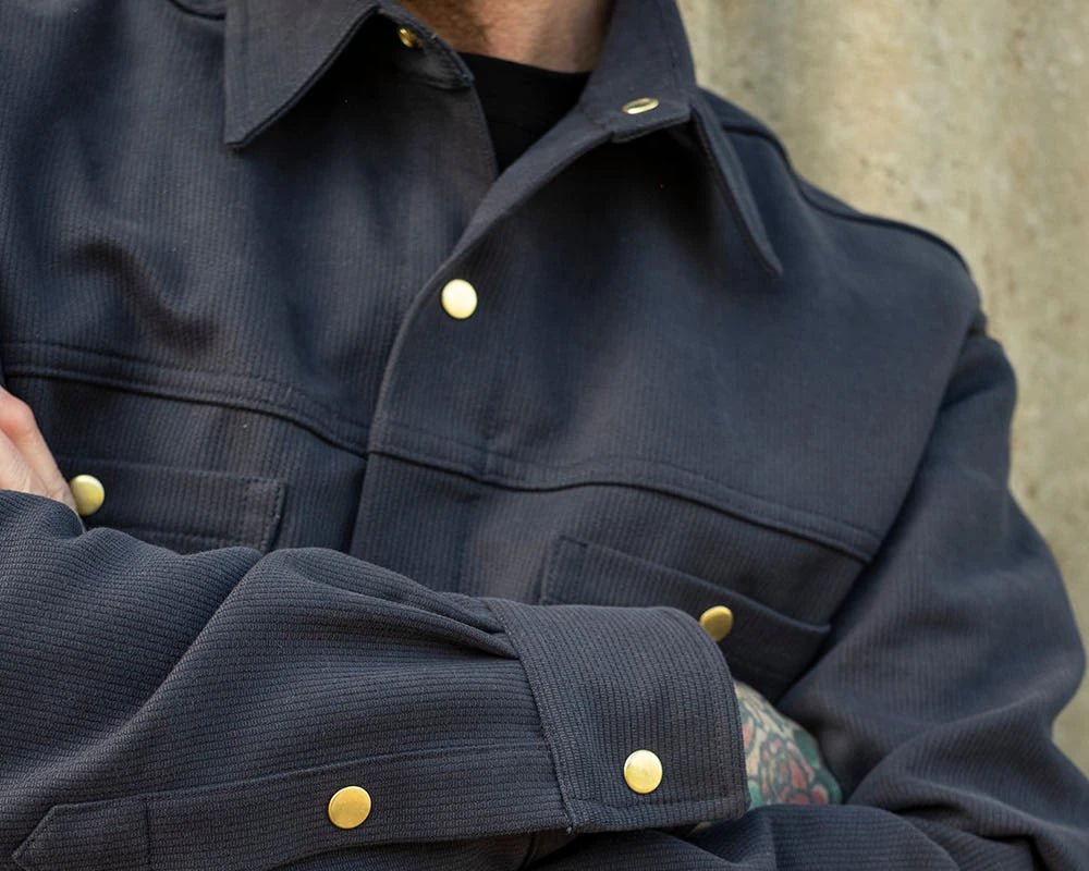 Close-up of a person wearing a dark navy corduroy overshirt with gold-tone snap buttons, with arms crossed against a textured concrete wall.