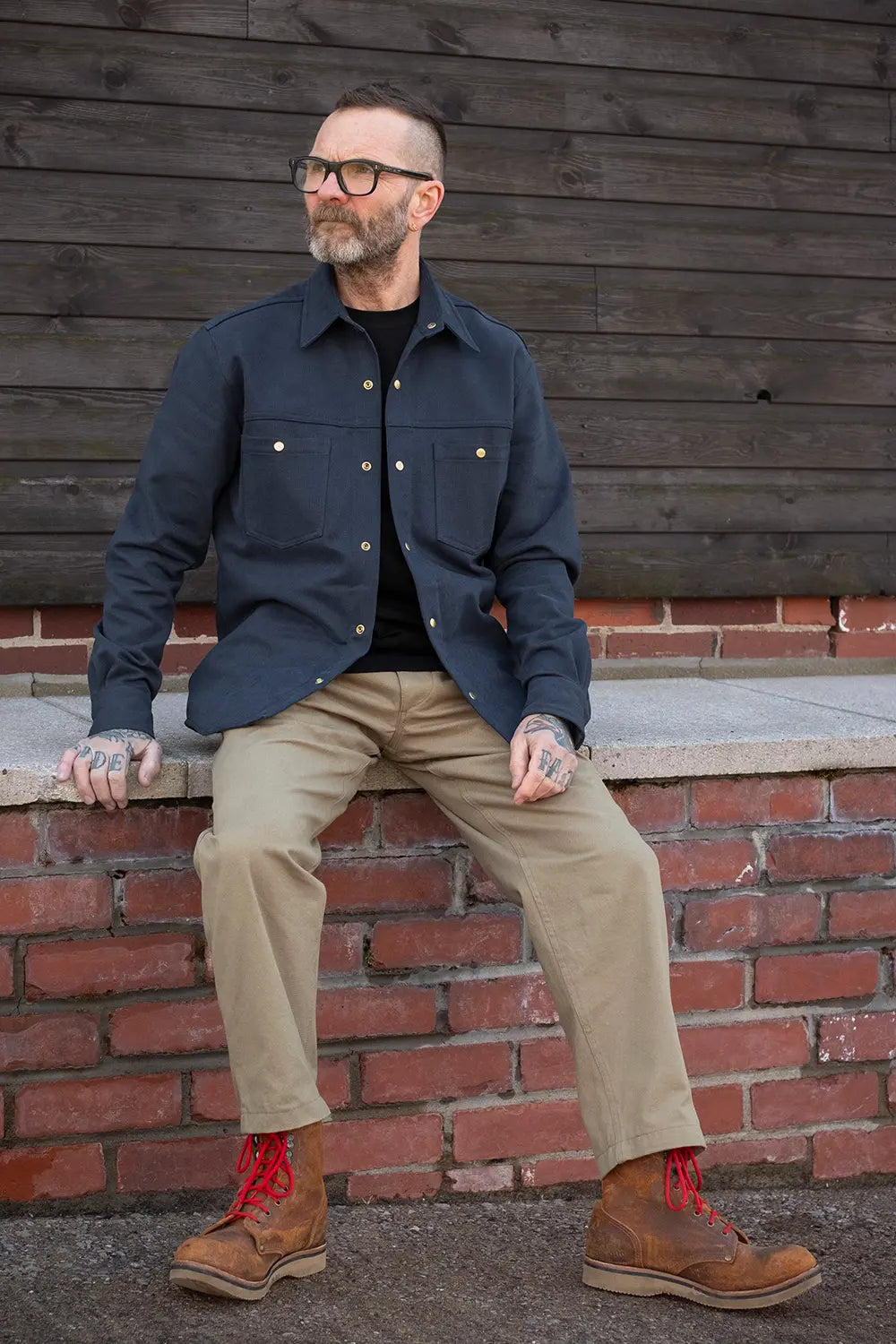 Ed wearing a bedford cord navy iron shirt, khaki pants, and brown boots with red laces sits on a brick ledge against a dark wooden wall.