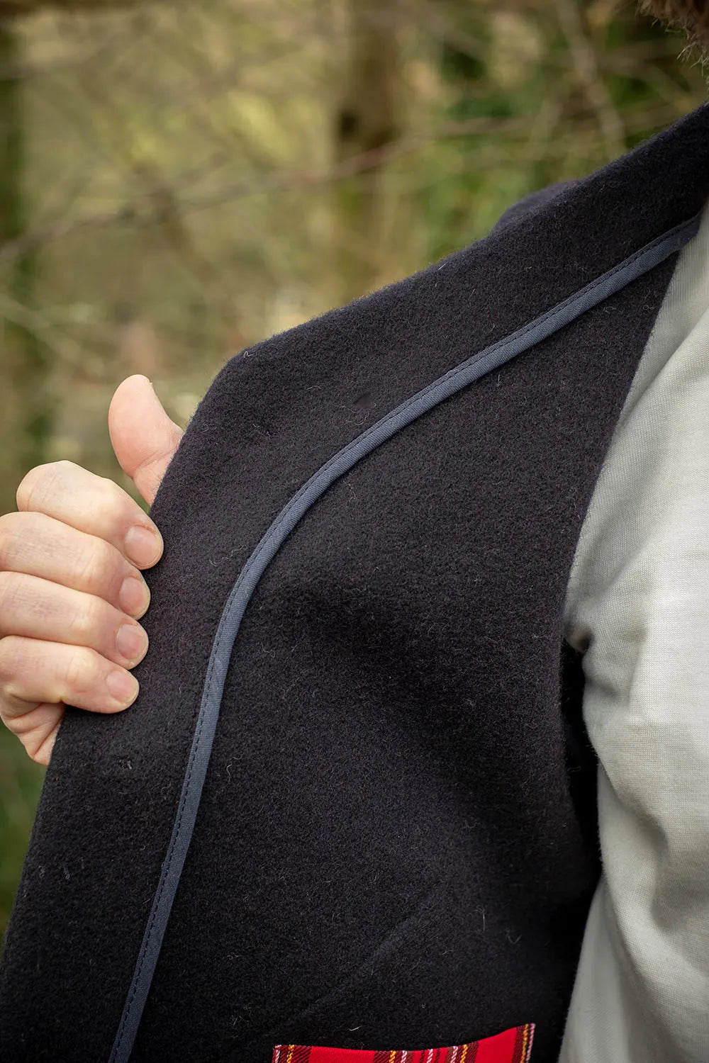 Close-up of a person wearing a navy wool vest with a hand holding the lapel, set against a blurred outdoor background.