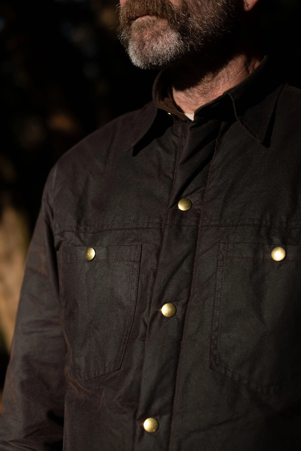 Close-up of a man wearing a dark brown waxed cotton button-up shirt with brass snap buttons and chest pockets.