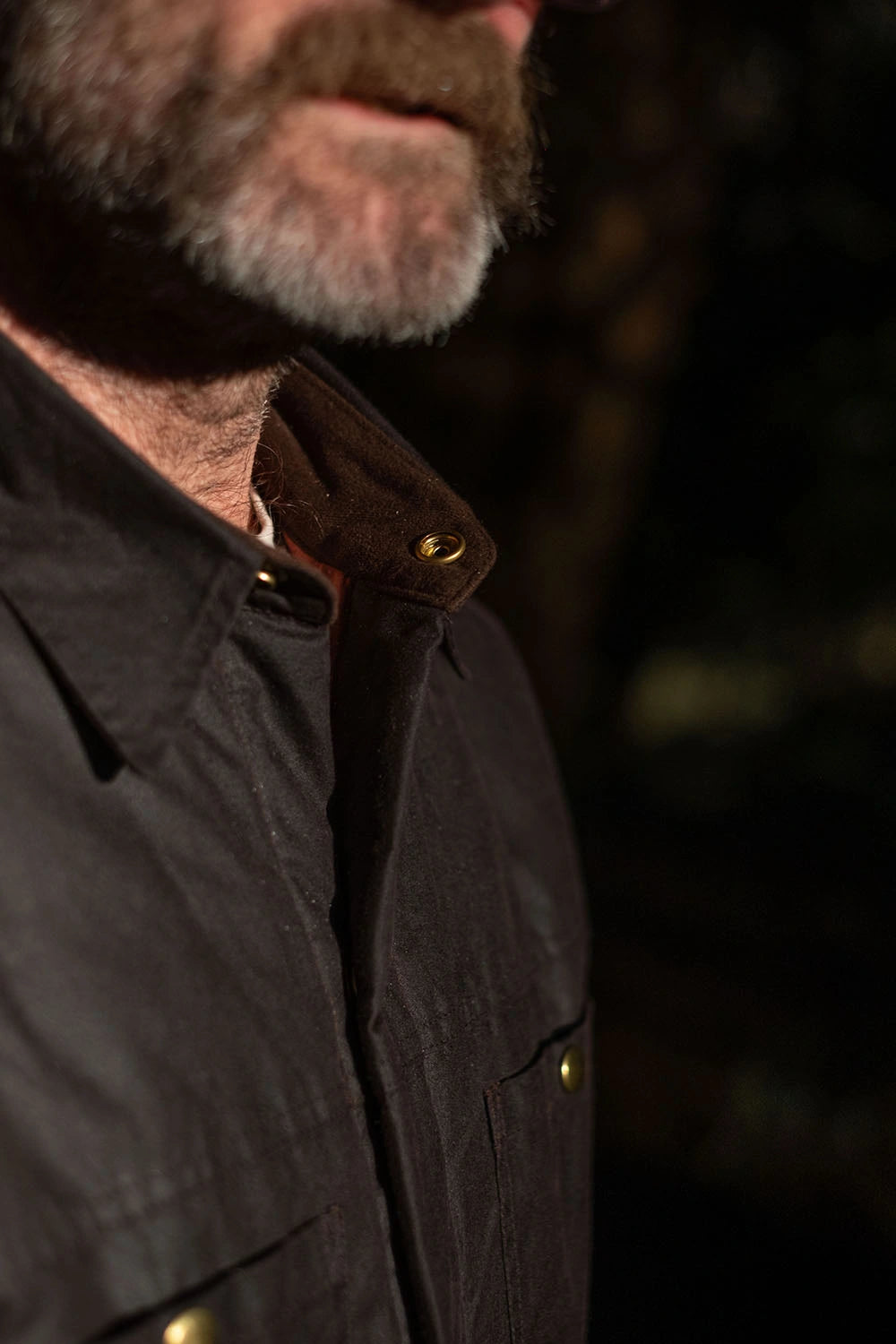 Close-up of a man wearing a dark brown waxed cotton overshirt with brass snap buttons and a brown corduroy collar, set against a dark, blurred background.