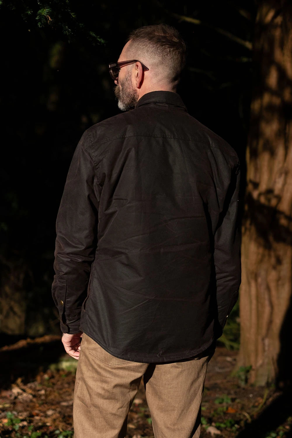 A man wearing a black long-sleeve button-up shirt and tan pants, photographed from behind in a dark outdoor forest setting.