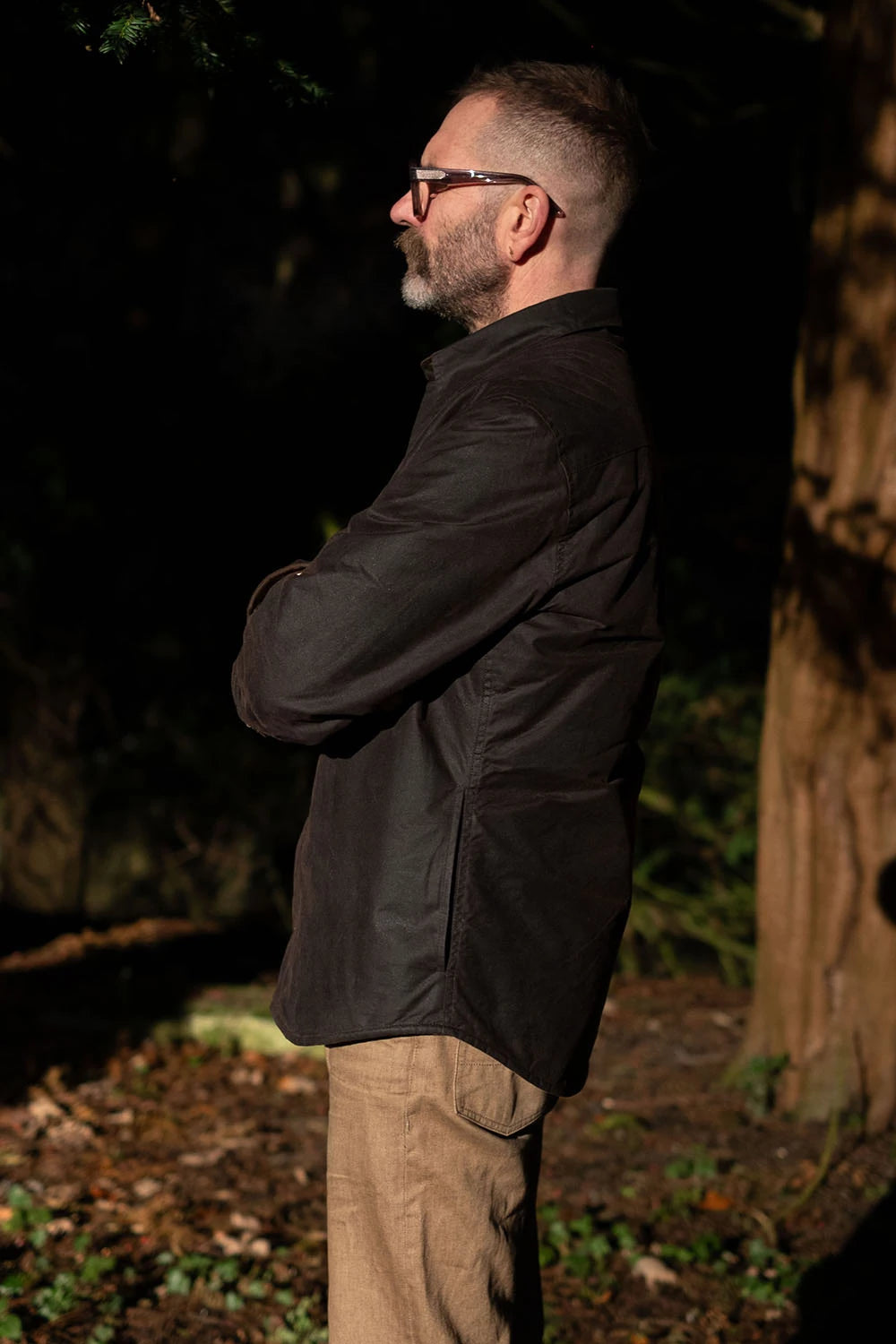 A man in profile wearing a dark jacket and khaki pants, standing outdoors in a wooded area at night with his arms crossed.