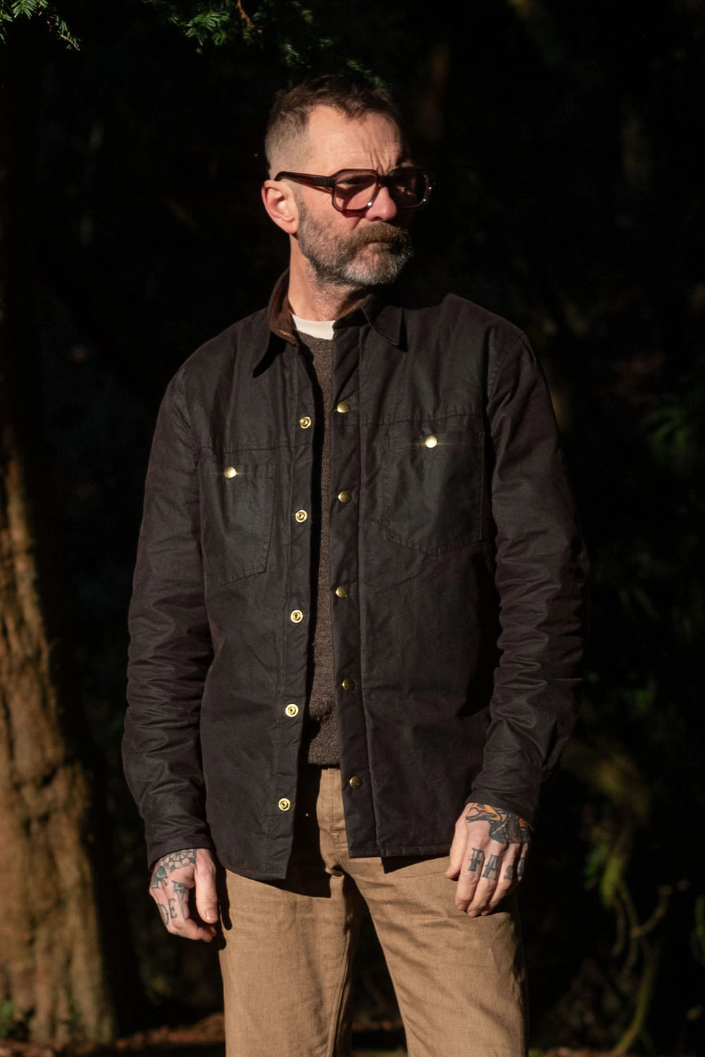 A man wearing a dark brown waxed cotton overshirt with brass buttons, tan pants, and glasses, standing outdoors in a dark, wooded setting.