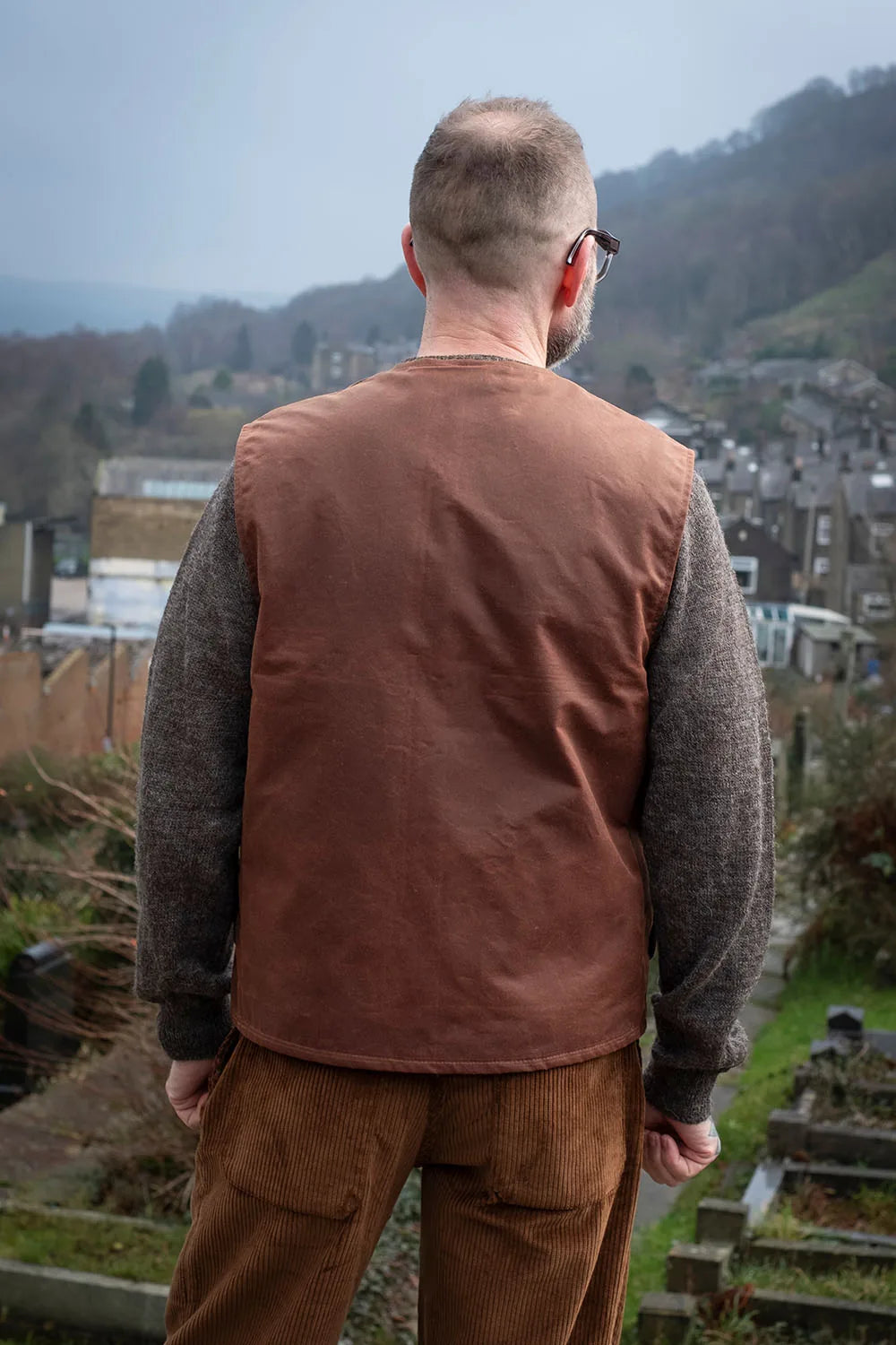 Ed viewed from behind wearing a Rascal vest over a gray knit jumper and brown corduroy trousers, standing outdoors with a village and hills in the background.