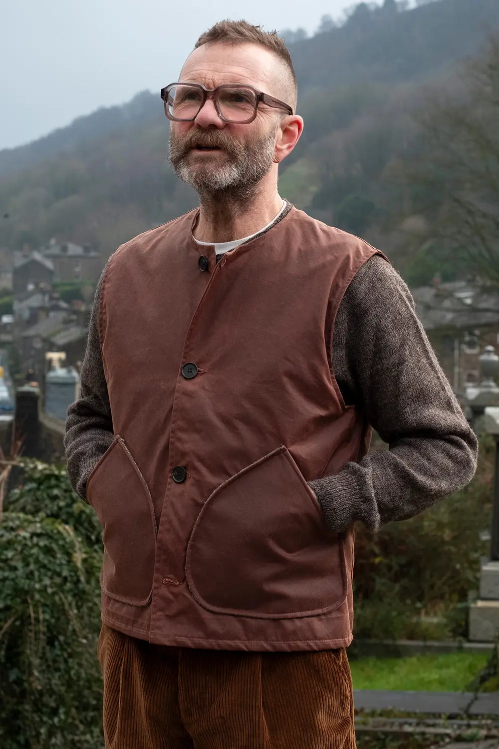 Ed wearing a rascal vest over a gray knit jumper, standing outdoors with a blurred background of hills and buildings.