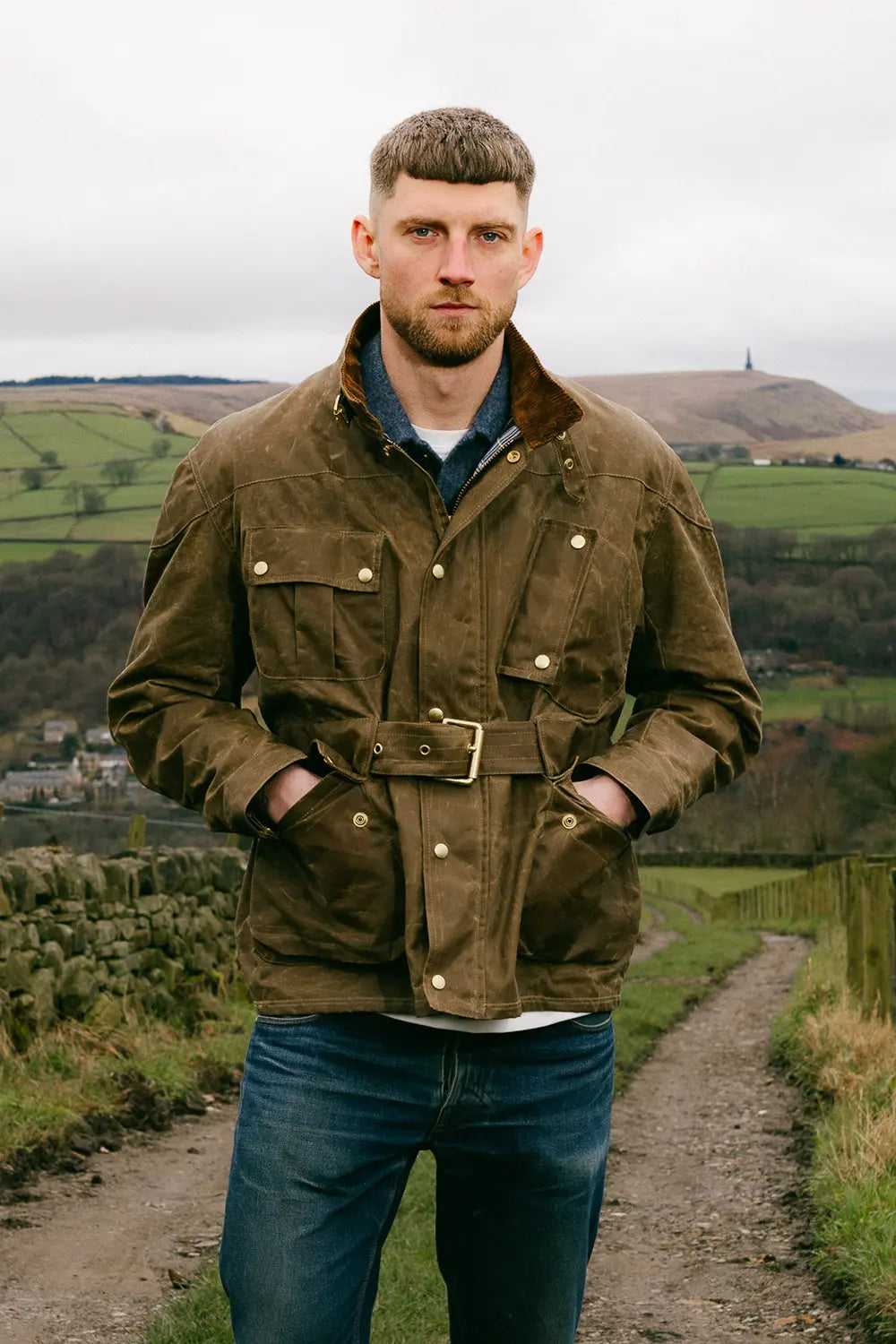 A man wearing a roadrunner jacket with a belted waist and brass buttons, standing on a dirt path with a rural landscape in the background.